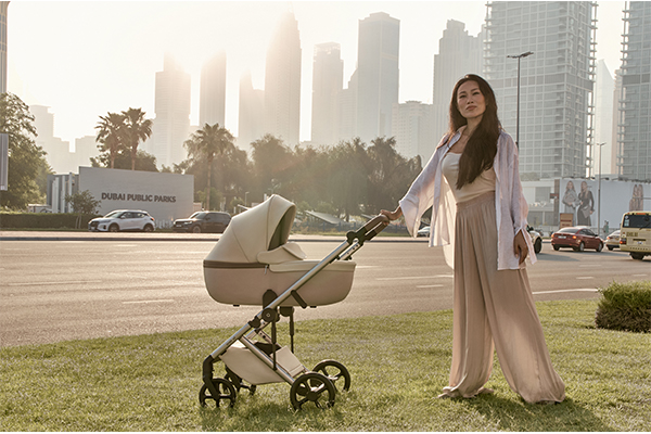 Kobieta spacerująca z wózkiem Anex Eli w miejskim otoczeniu; beżowy wózek 2w1 z gondolą, elegancki i komfortowy model idealny do codziennych spacerów po mieście.